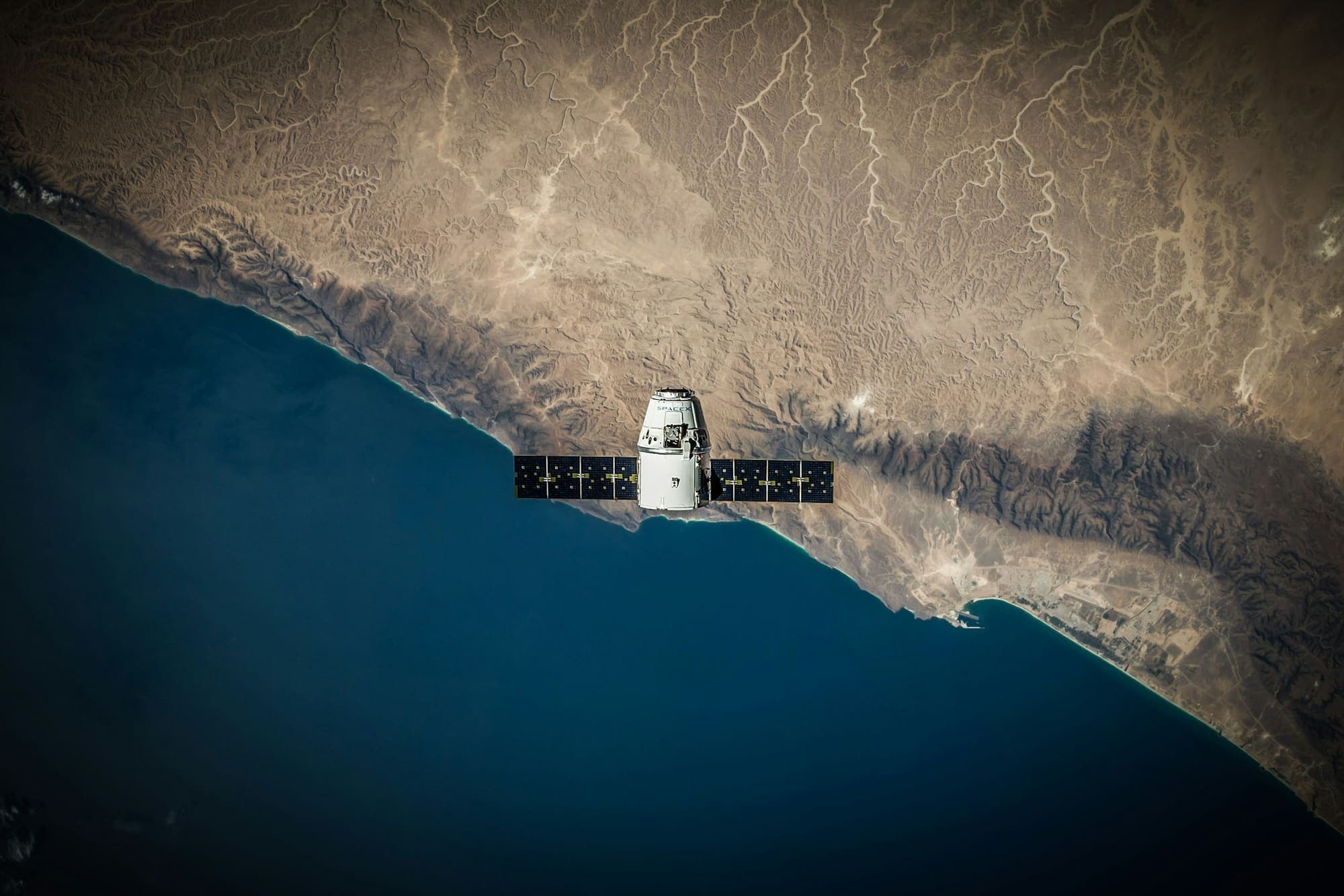Spacecraft with solar panels orbiting above a desert coastline